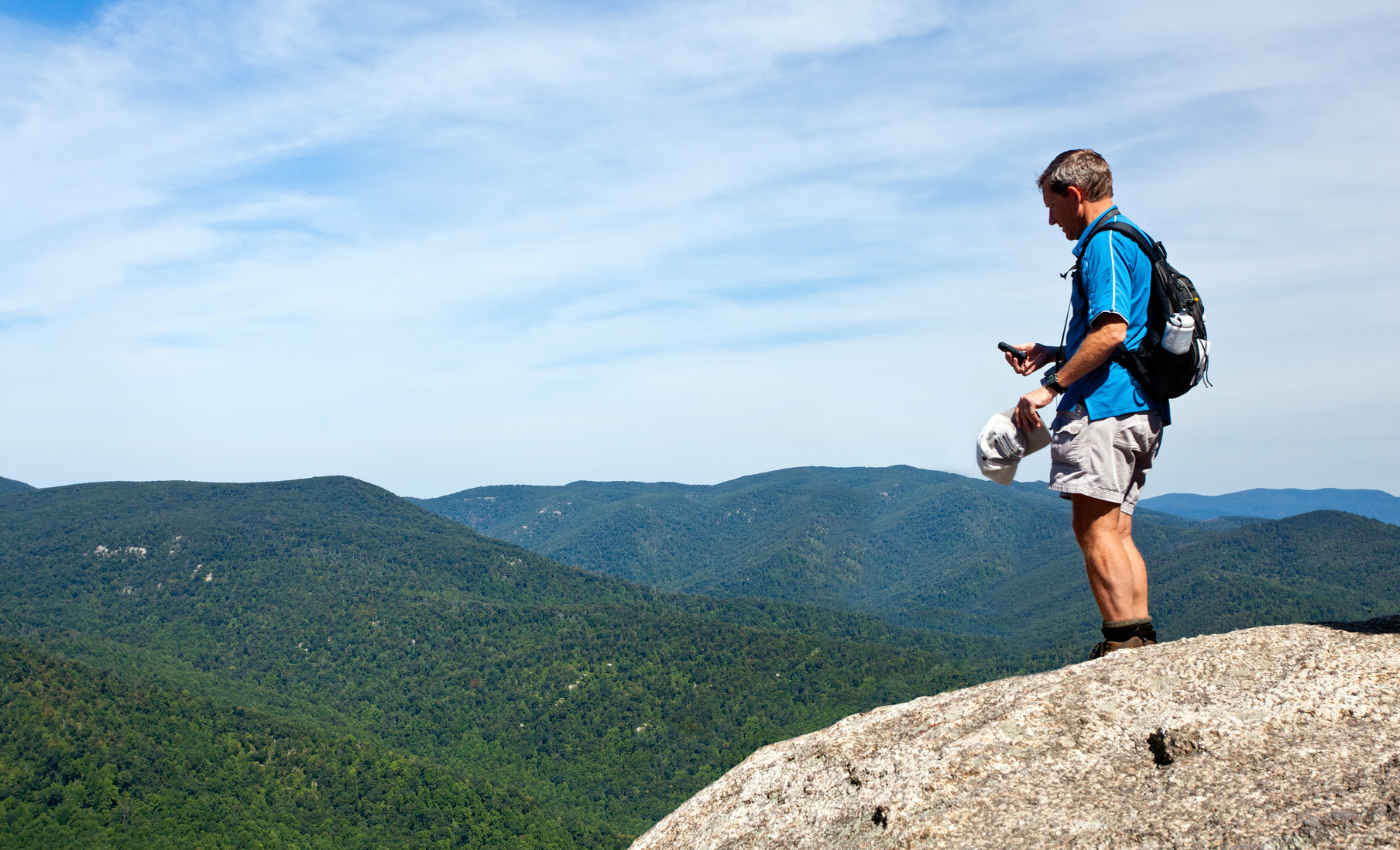 a man stands on a rock on the peak of a mountain with rolling green mountains in the background