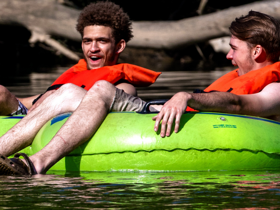 two men sit on green inner tubes and float a river