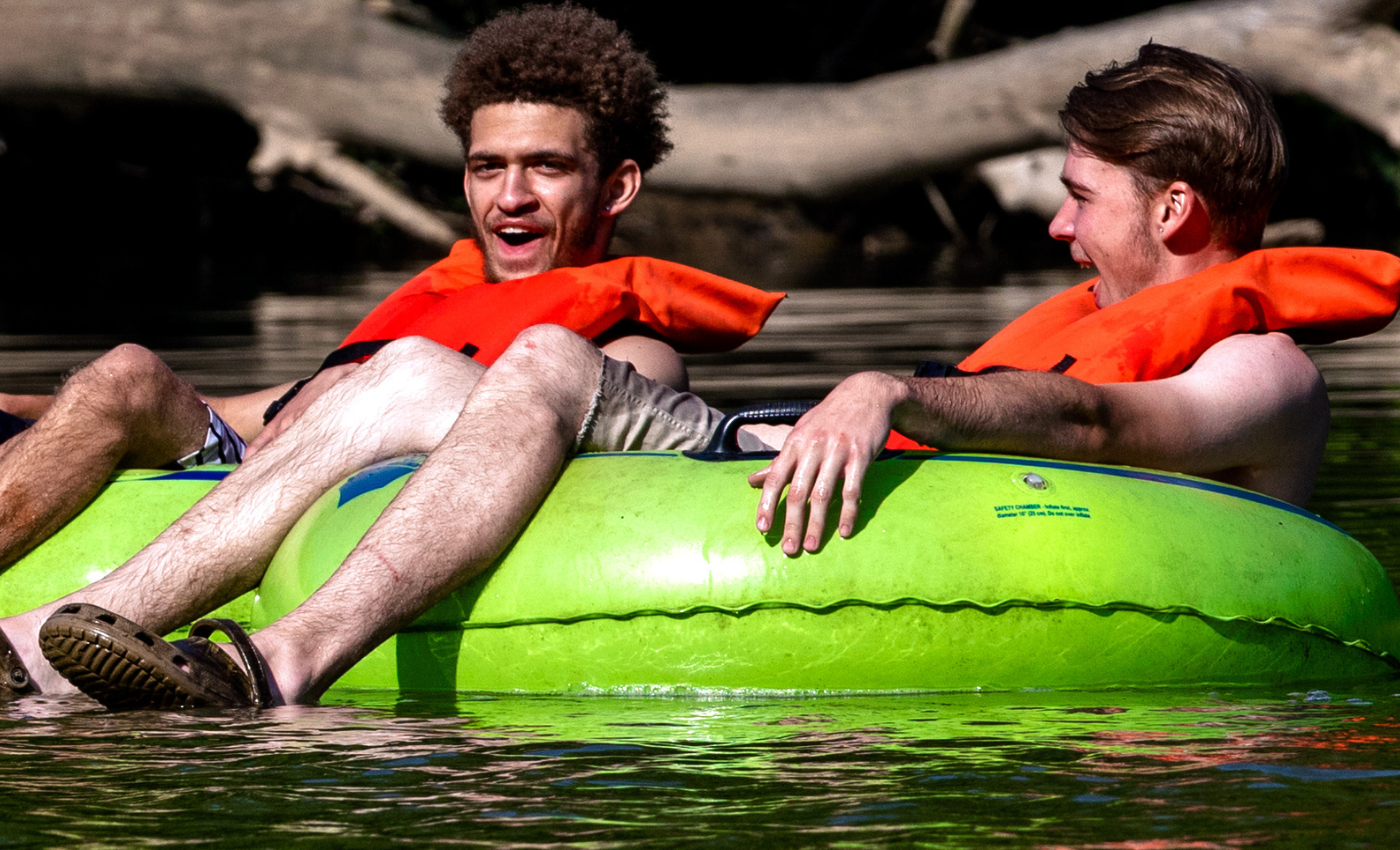 two men sit on green inner tubes and float a river